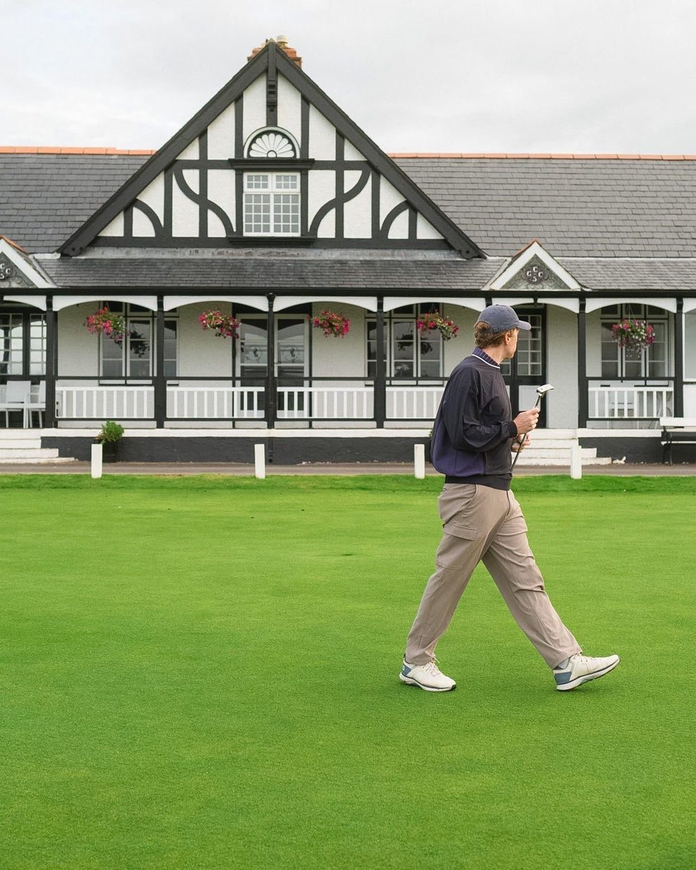 A golfer in a navy Windcrew walking past a black-and-white Tudor clubhouse