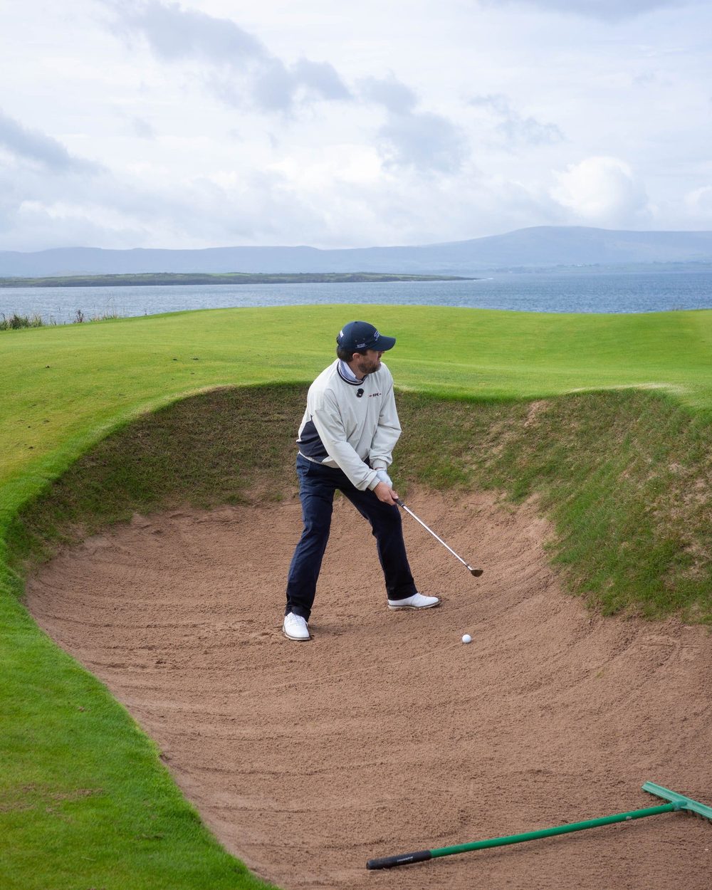 A golfer in a stone Windcrew playing out of a pot bunker with the sea behind