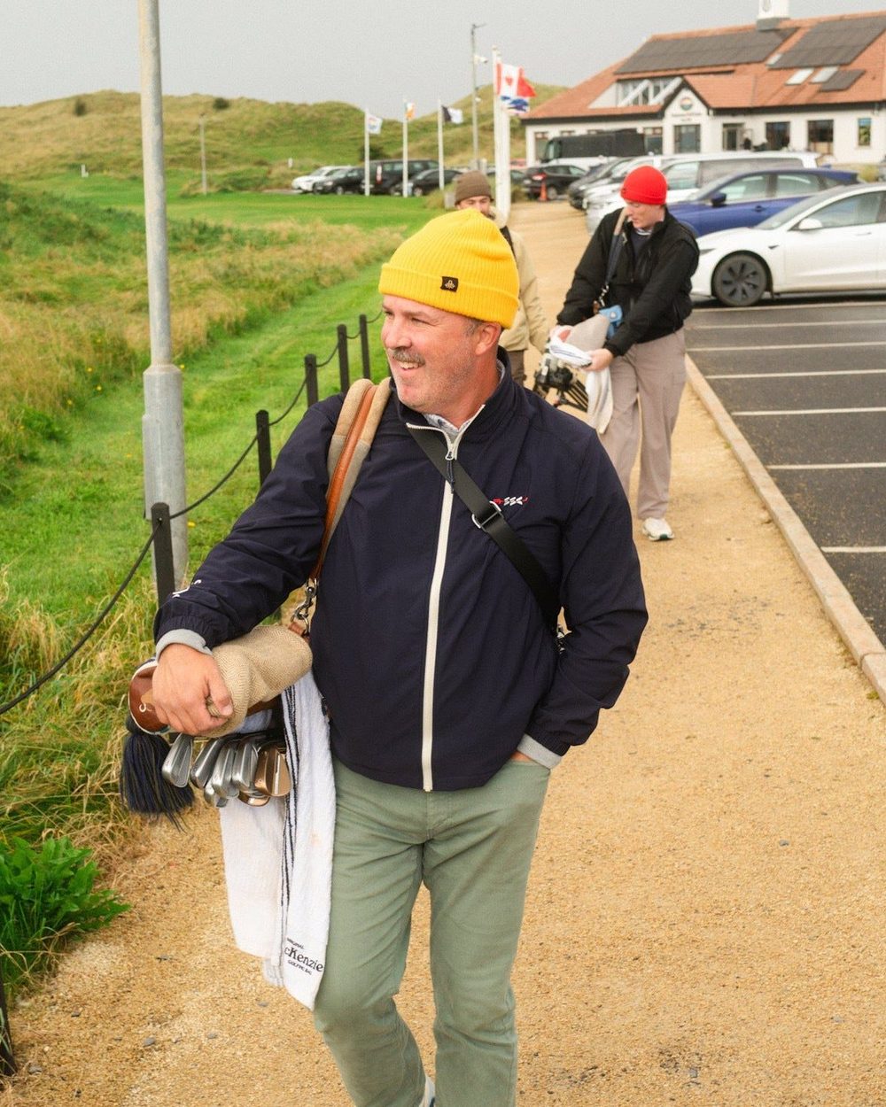 A smiling golfer in a navy Tech Jacket and yellow beanie carrying his bag by the car park