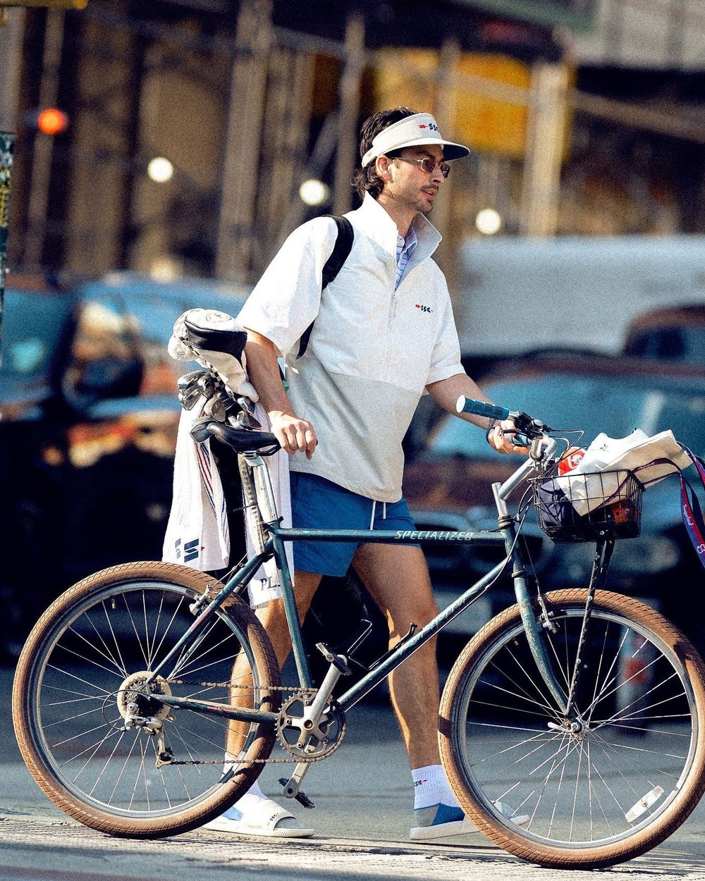 A man in a Windshirt riding a bicycle with a golf bag through a city street