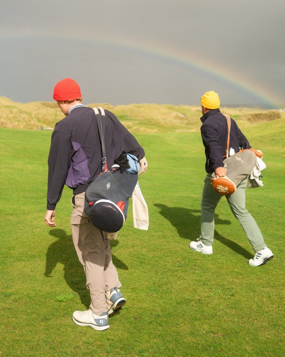 Two golfers in red and yellow beanies carrying bags across an Irish links under a rainbow