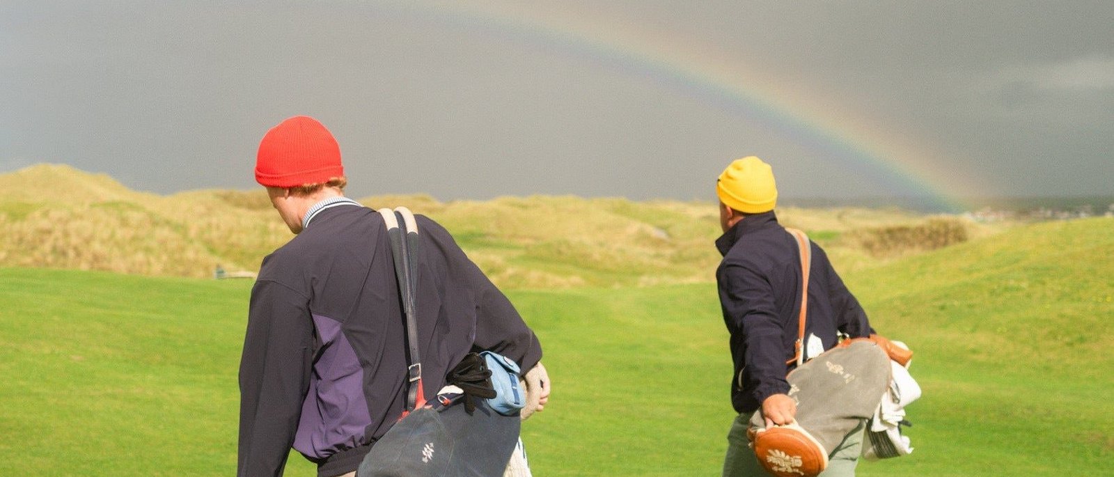 Two golfers in red and yellow beanies carrying bags across a links course in Ireland under a rainbow