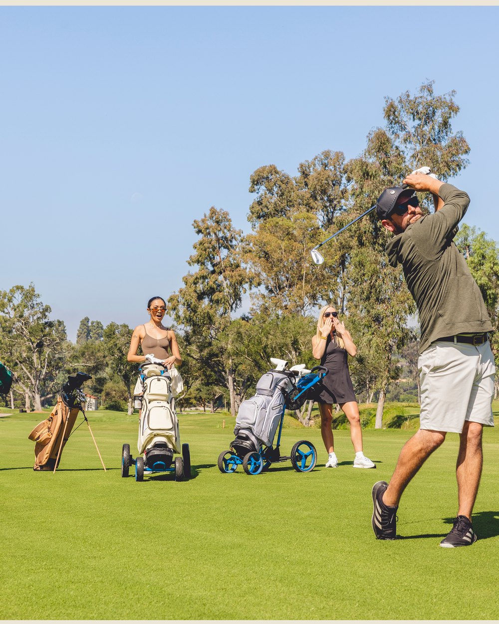 Three golfers on a sunny fairway with two Sunday Golf bags on push carts, one golfer finishing his swing