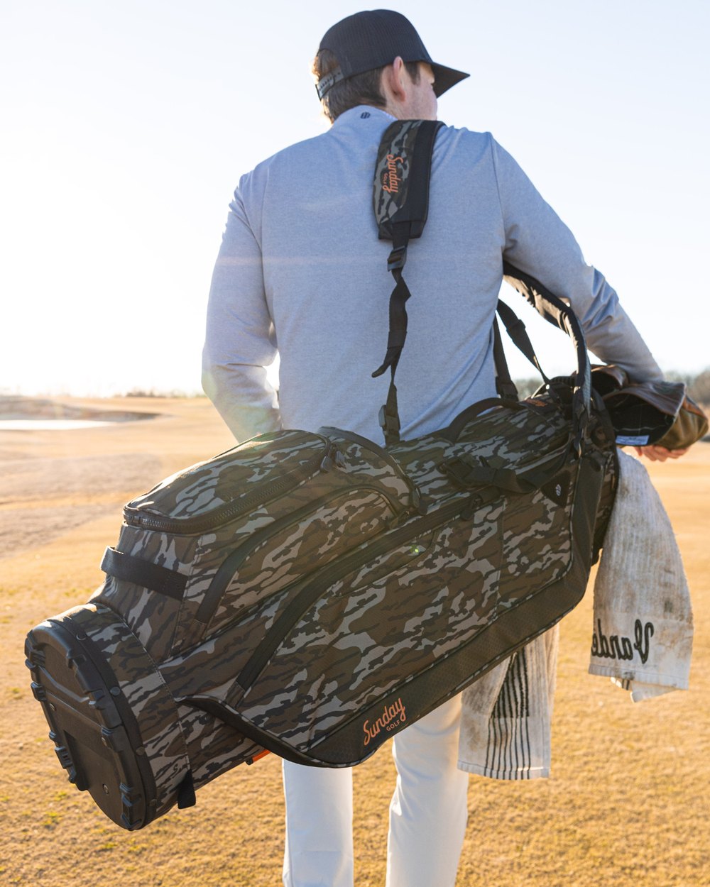 A golfer carrying a camo Sunday Golf stand bag over his shoulder with a towel clipped on