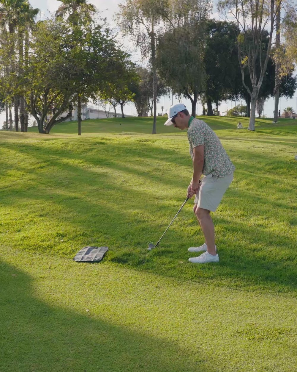 A golfer in shorts chipping from a grassy slope among palm trees