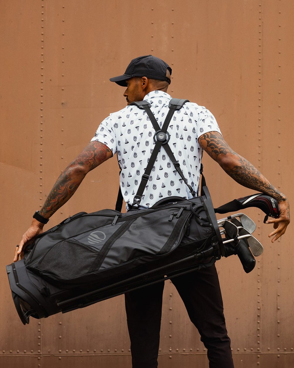 A man carrying a black Sunday Golf stand bag on a double strap in front of a rusted wall