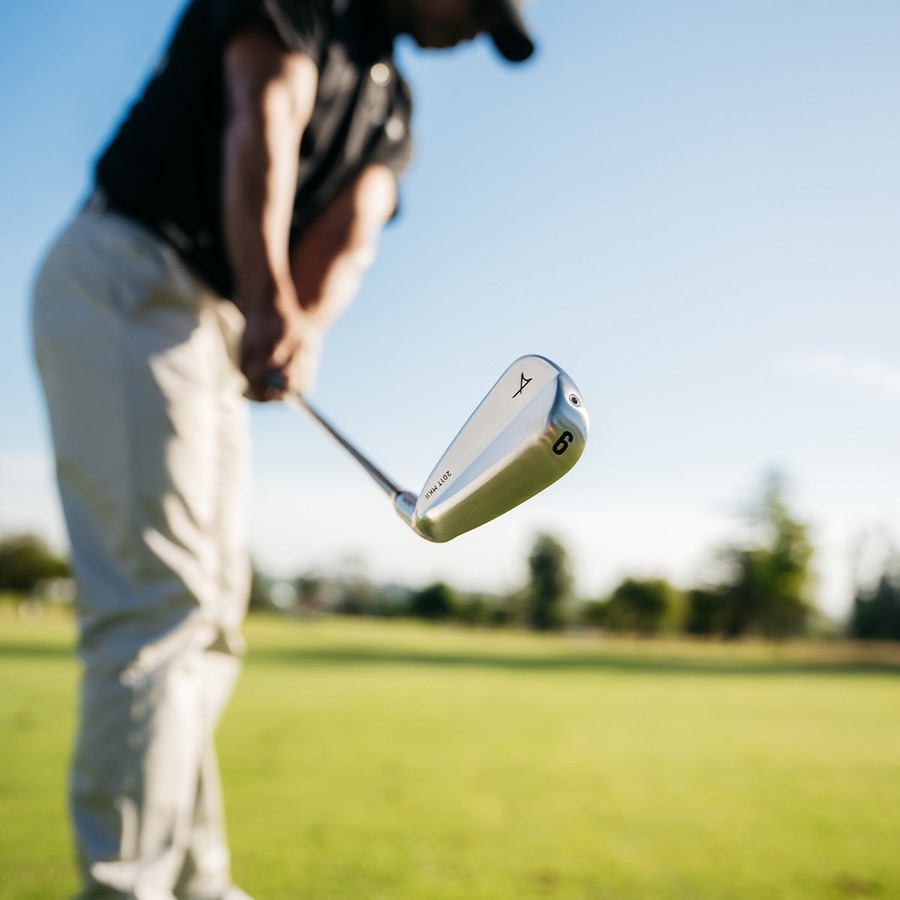 A golfer addressing a ball on the fairway with a Takomo iron, tree line behind