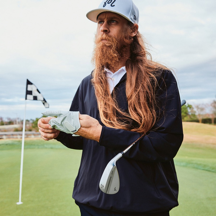 A bearded golfer in a navy jacket standing on a green holding the Skyforger 002 wedge, flagstick behind