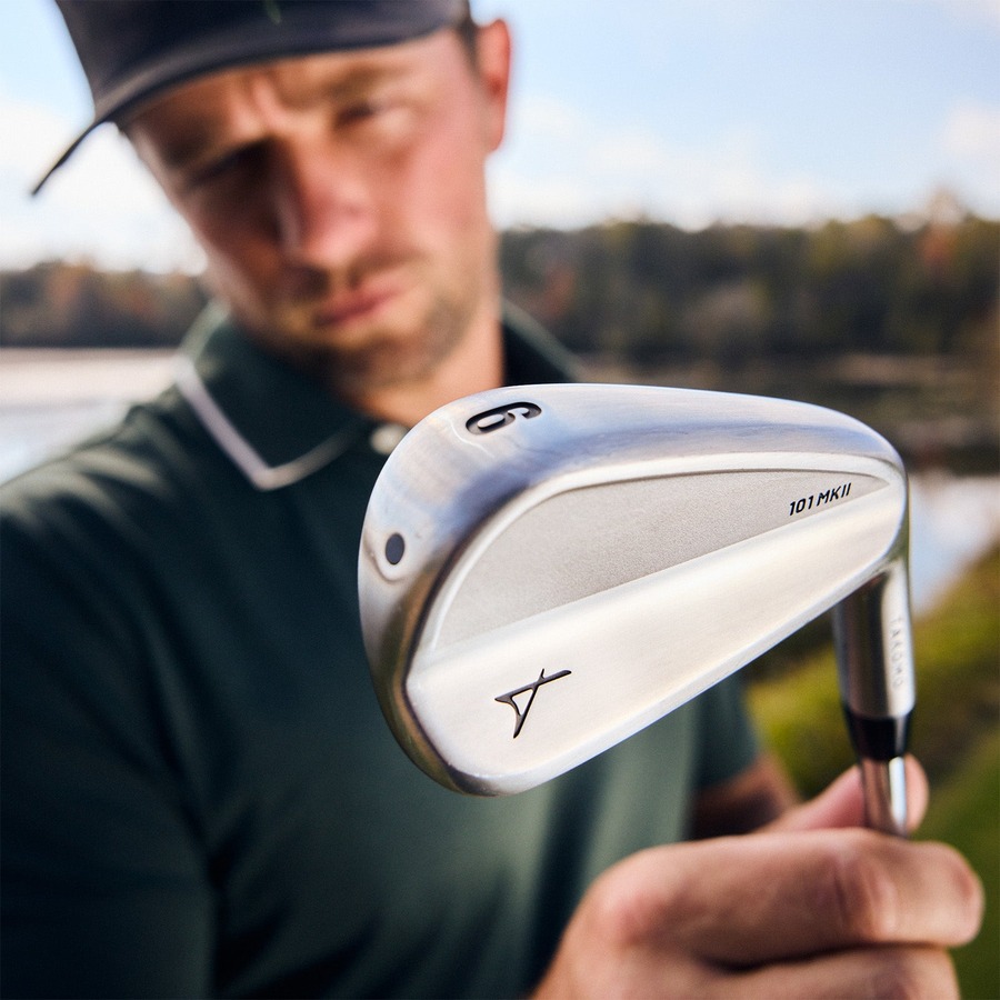 A golfer in a cap and dark polo holding a Takomo 101 MKII iron, open course behind