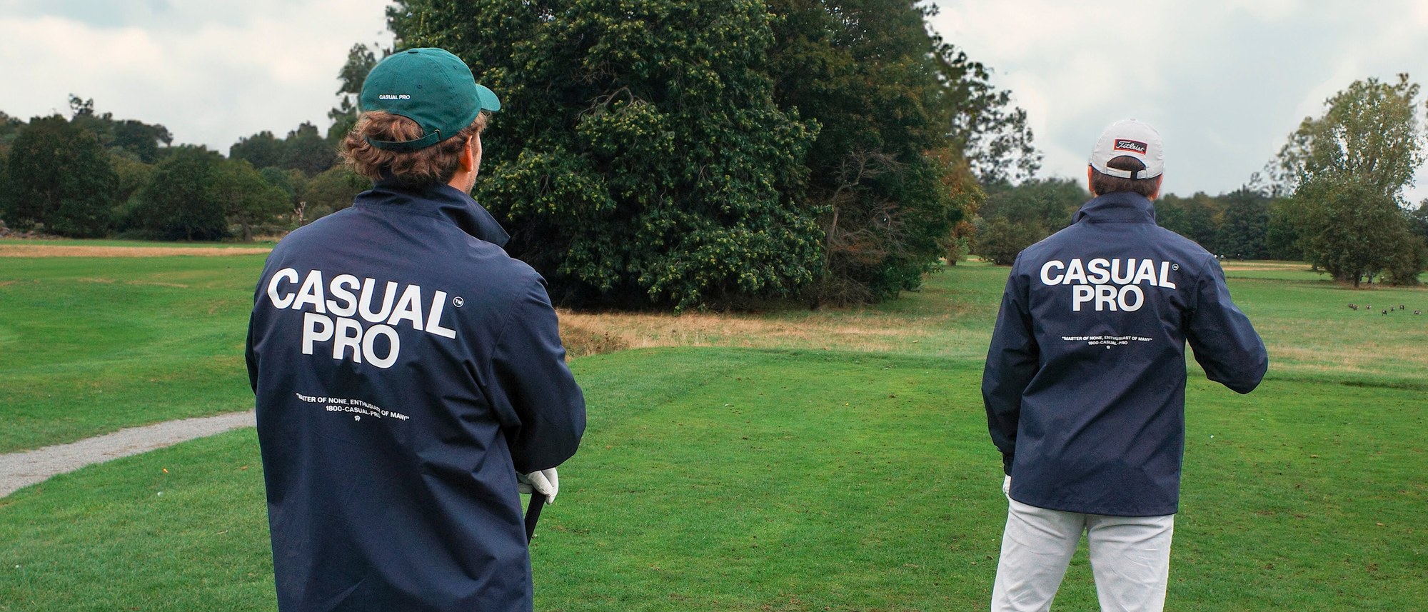 Two golfers seen from behind on a fairway wearing navy Casualist windbreaker coach jackets with CASUAL PRO printed on the back