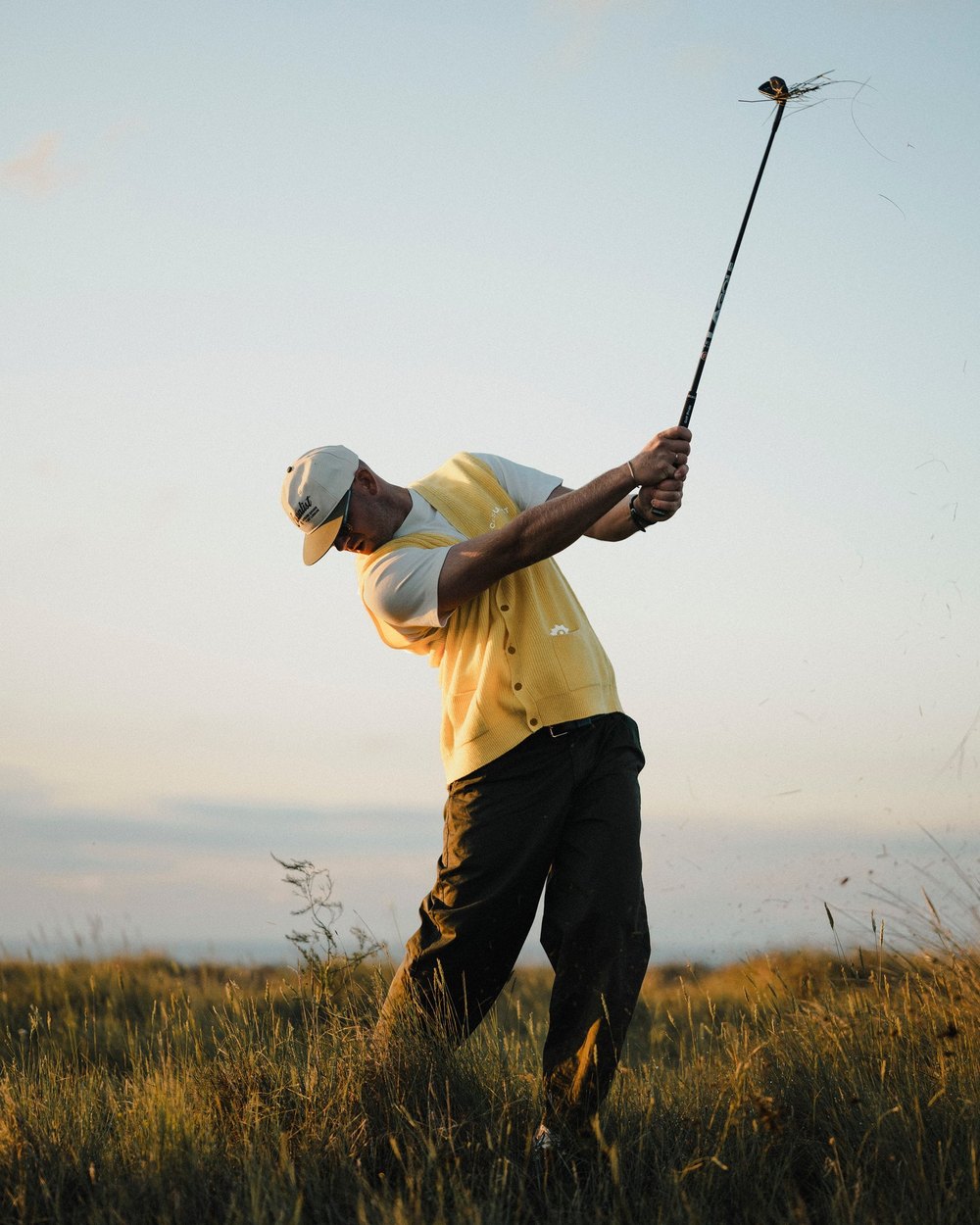 A golfer in a yellow Casualist cardigan vest mid-swing in long grass