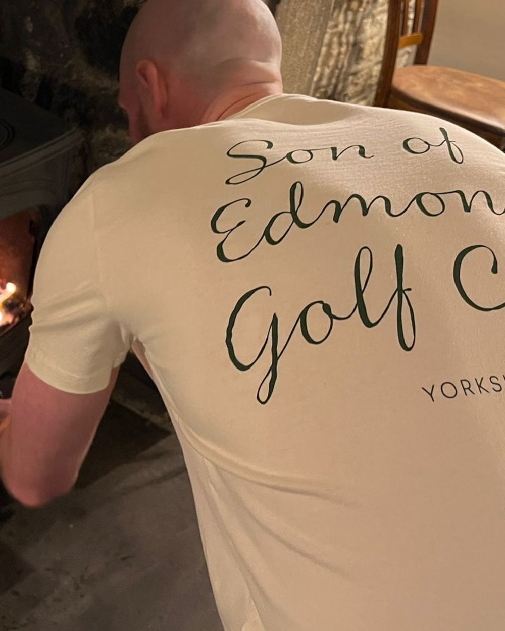 A man by an open fire in a Son of Edmond Golf Club T-shirt