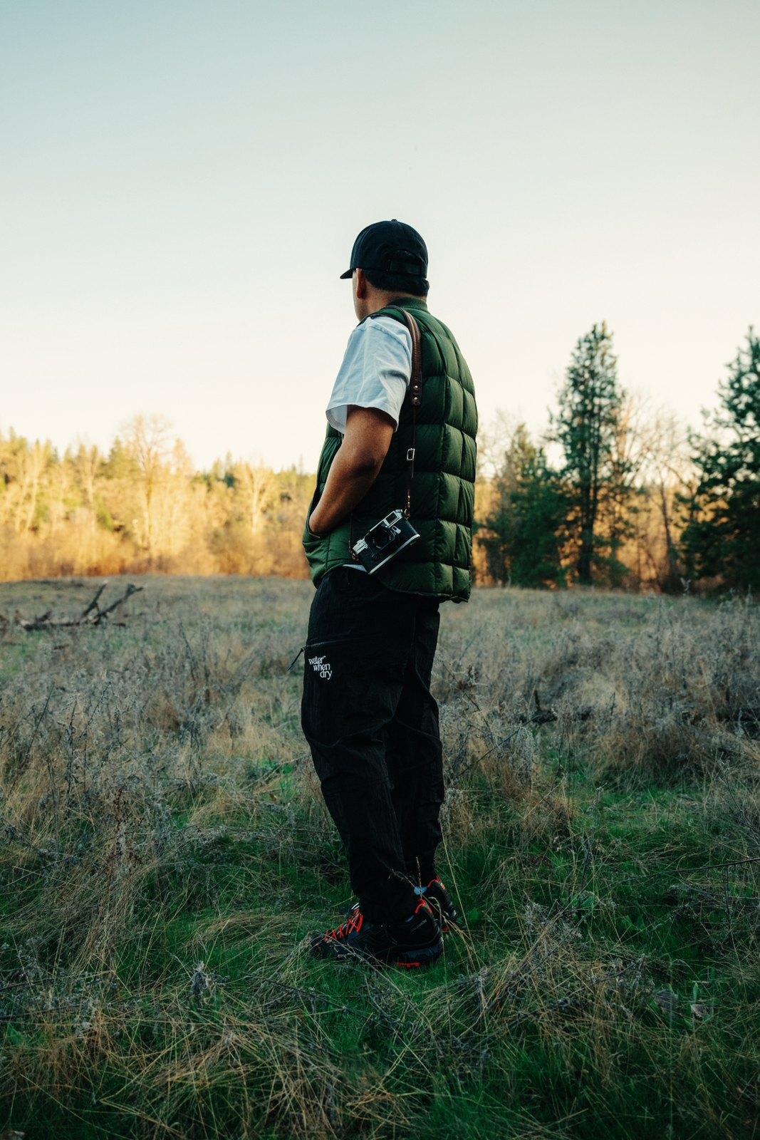 A man in a green puffer vest and black utility pants standing in a field at sunset