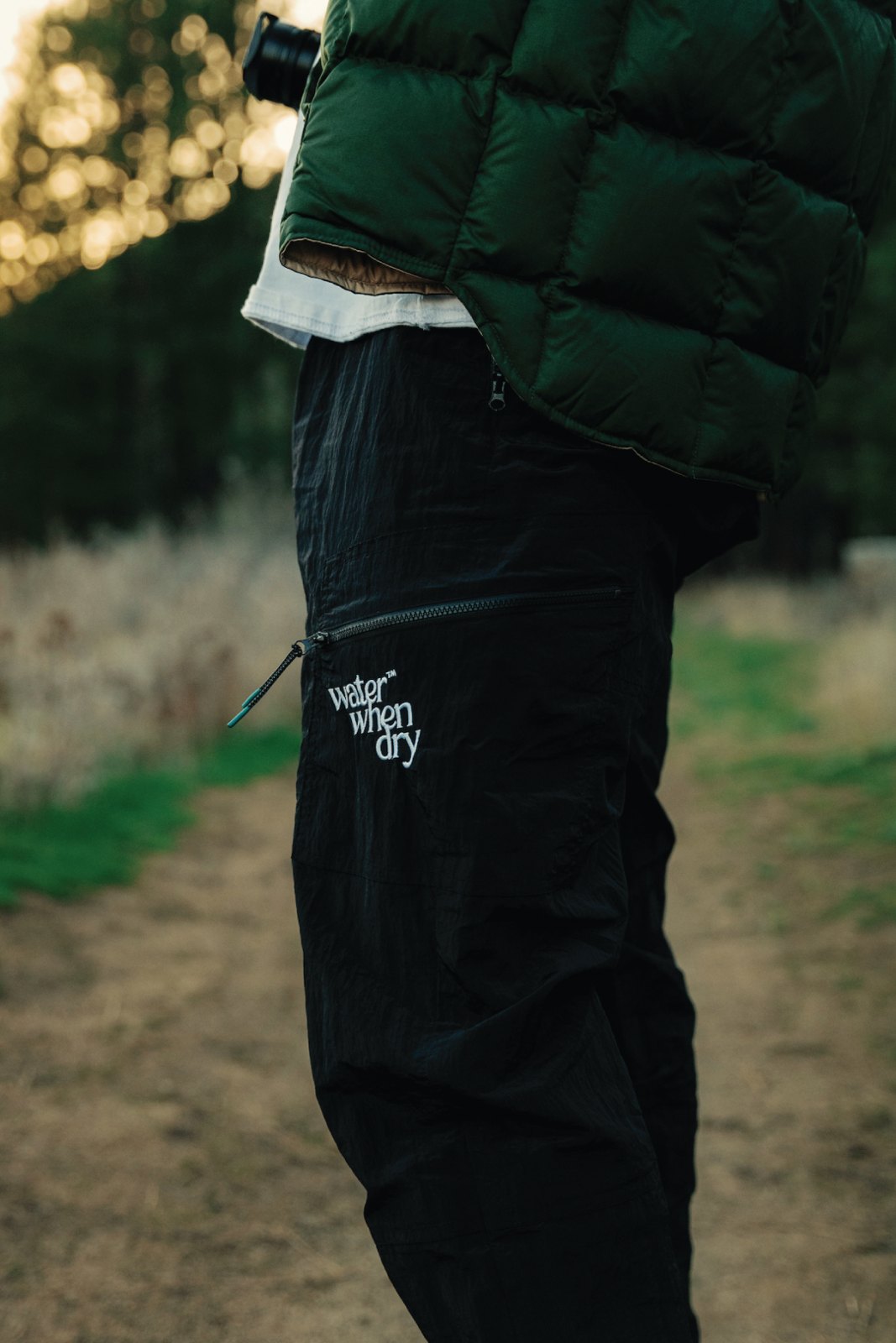 Close-up of black utility pants with the script logo at the knee on a dirt path