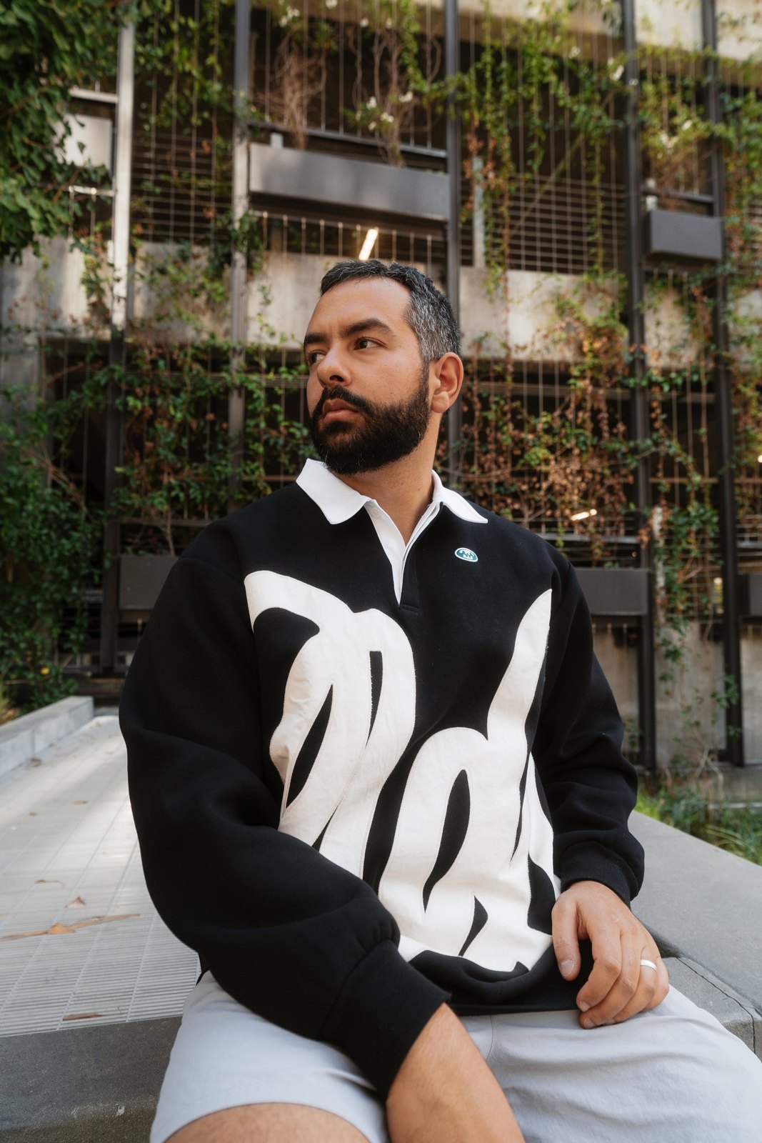 A man in the black pullover sitting on a concrete ledge in front of a wall of vines