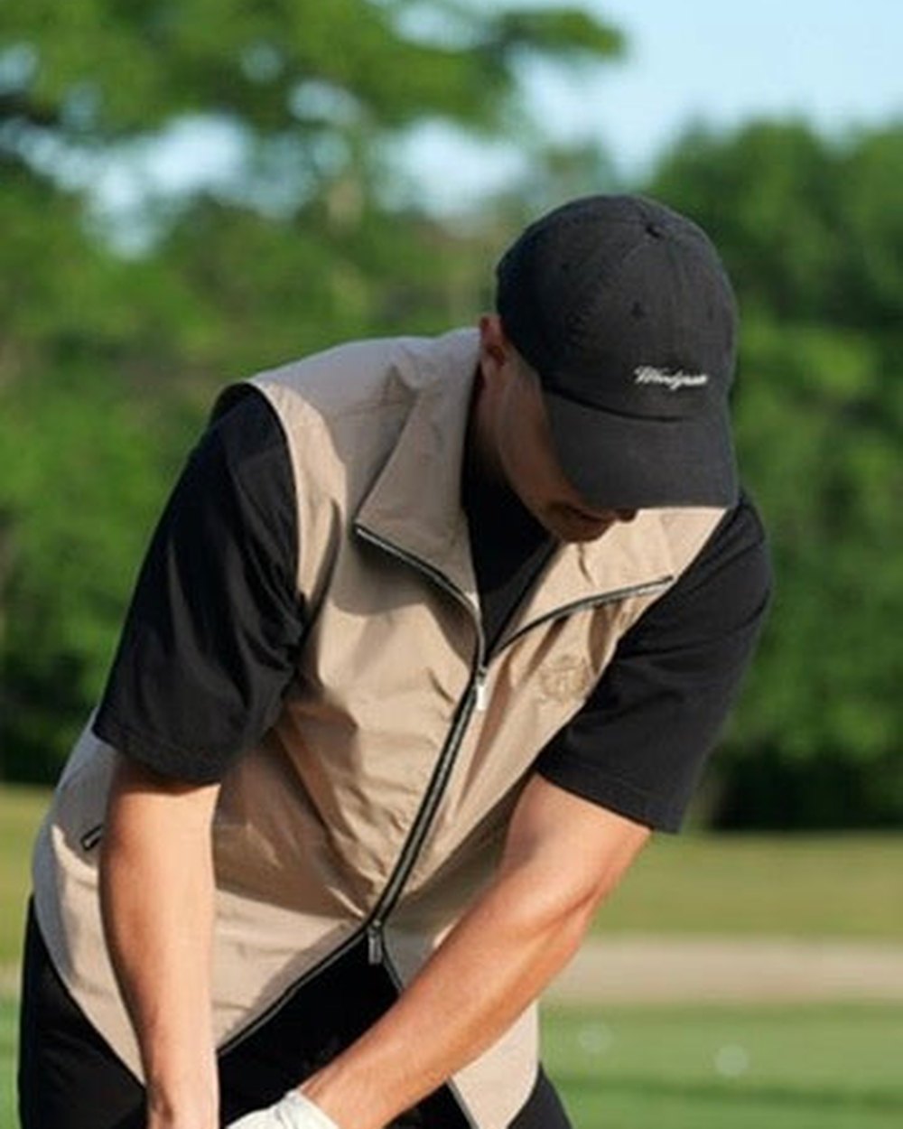A golfer in a black Woodgrain script cap and tan vest mid-swing